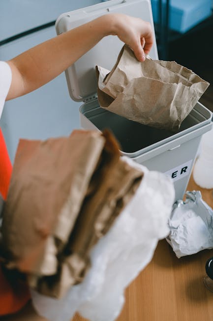 A person placing a crumpled brown paper bag into a small, grey static rubbish bin with a black interior, which is situated on a wooden surface near a wall. The individual's arm is visible, holding the